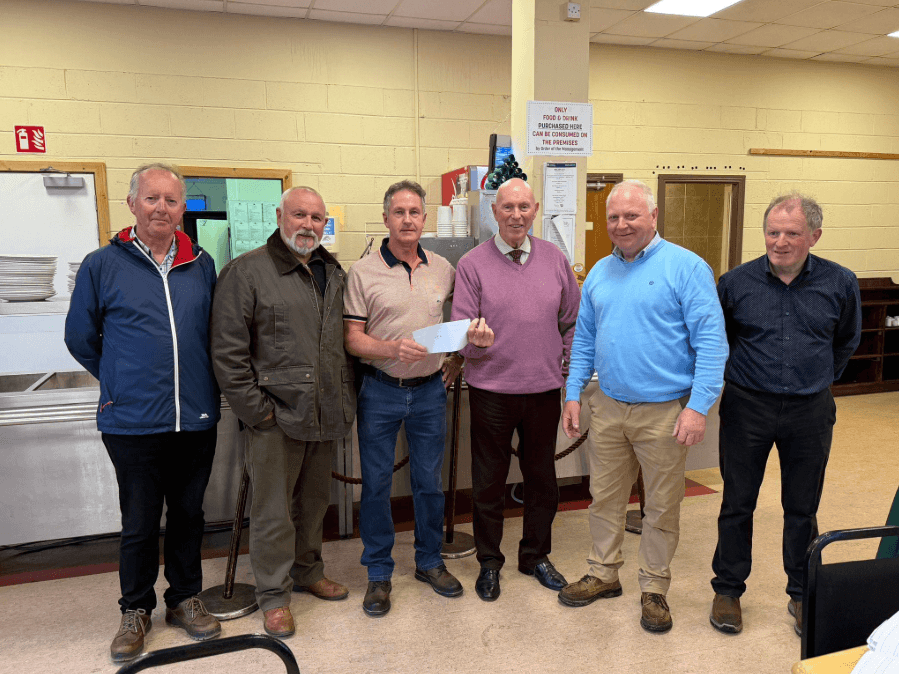 Six men standing next to each other, faced towards the camera, and two men in the middle holding the cheque