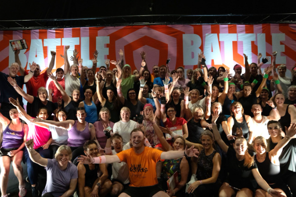 A large group of participants celebrating together at an indoor fitness event, smiling, cheering, and posing in front of the event backdrop after a high-energy group session.
